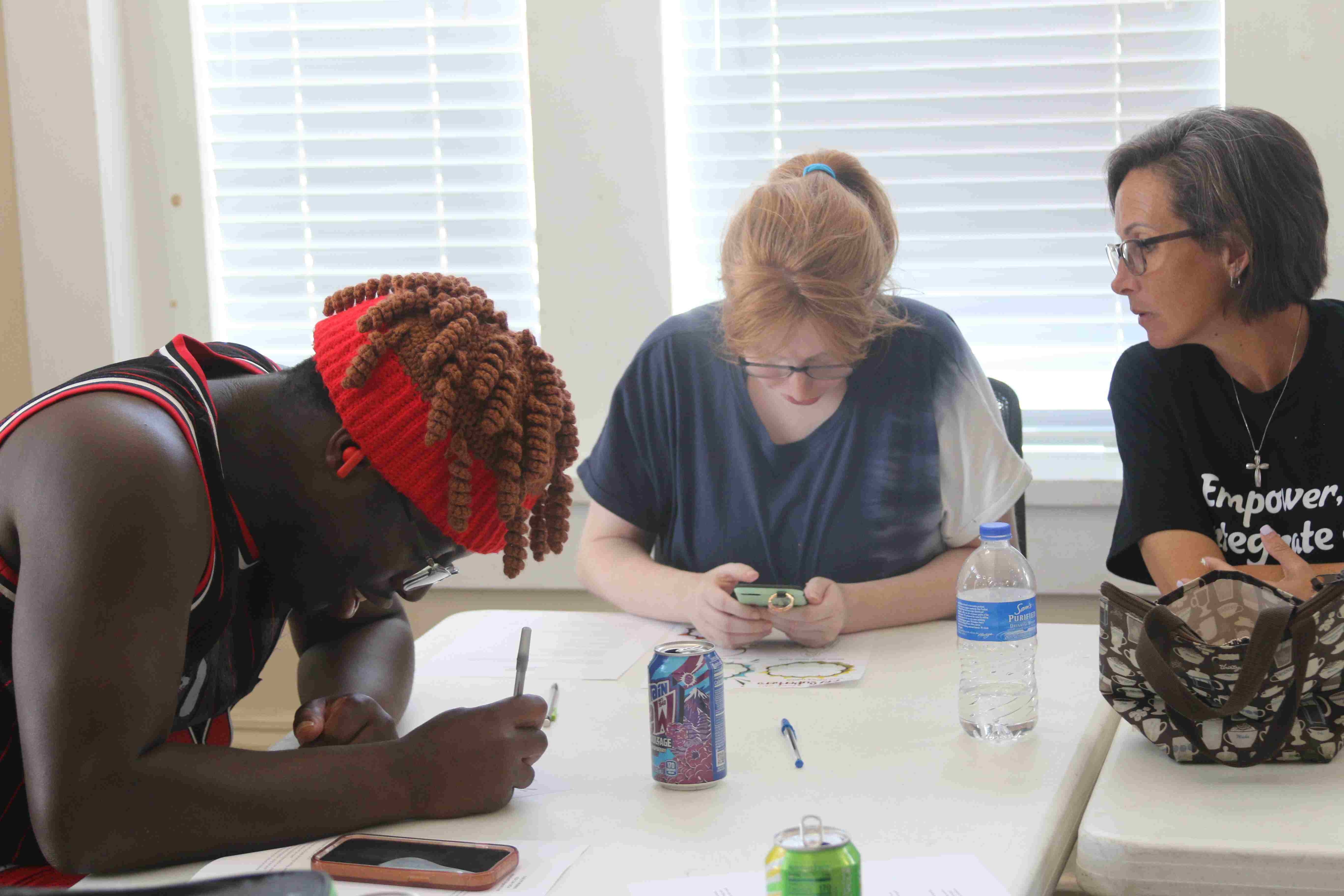 three people seated at a table. two are looking at a phone. one is drawing.